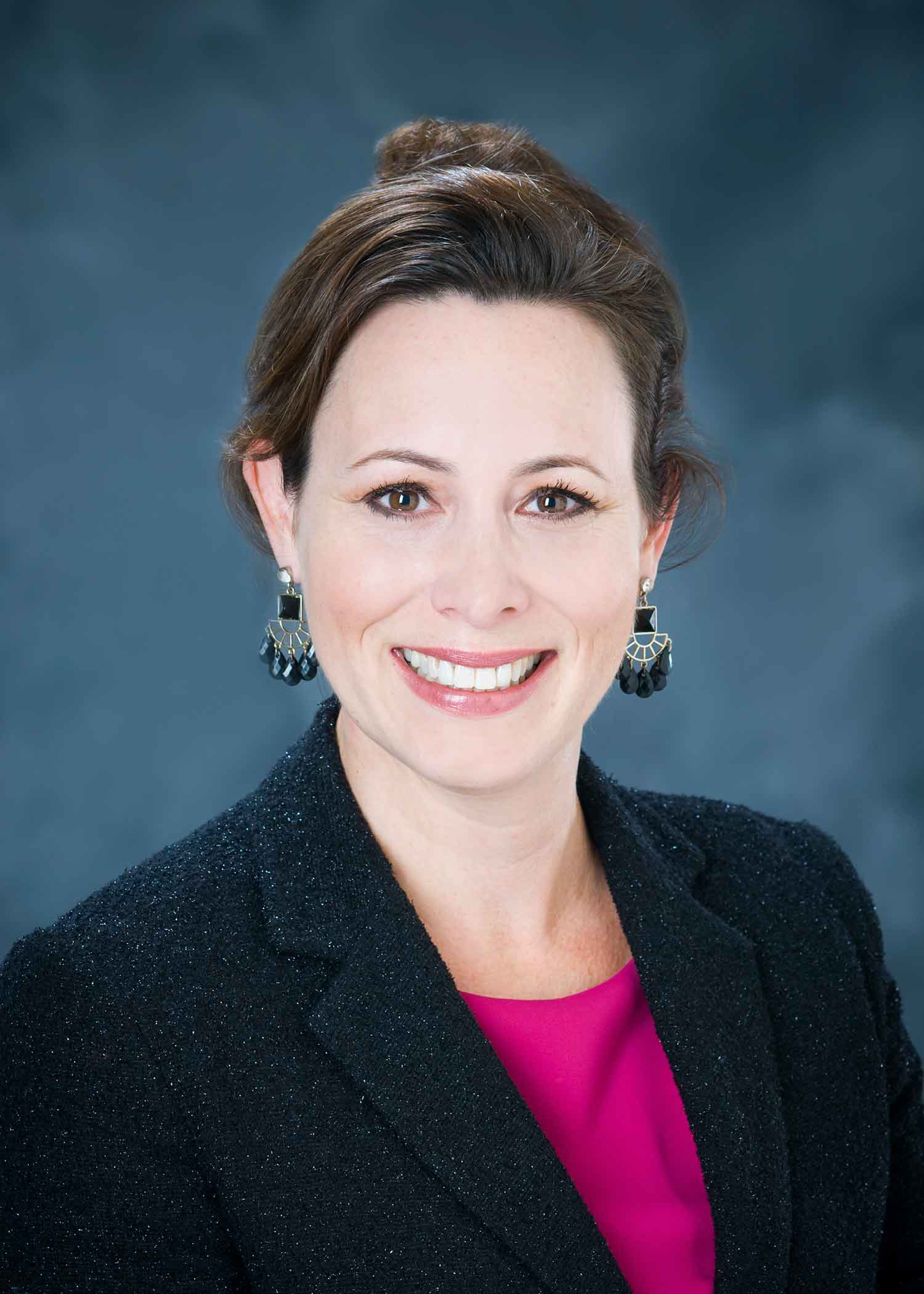 Professional photo of Christa Haney, wearing a pink blouse and dark blazer against a blue background.