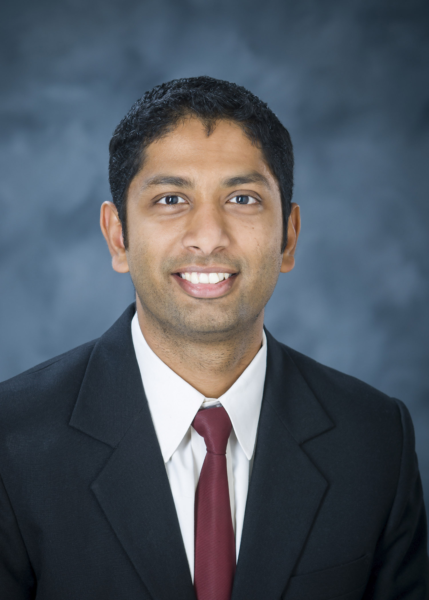 Professional photo of Varun Paul, wearing a black suit and red tie against a blue background.