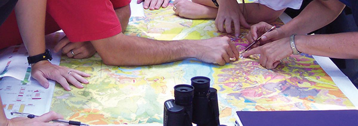  Students working on a map exercise; multiple hands pointing out a location on a colorful map. 