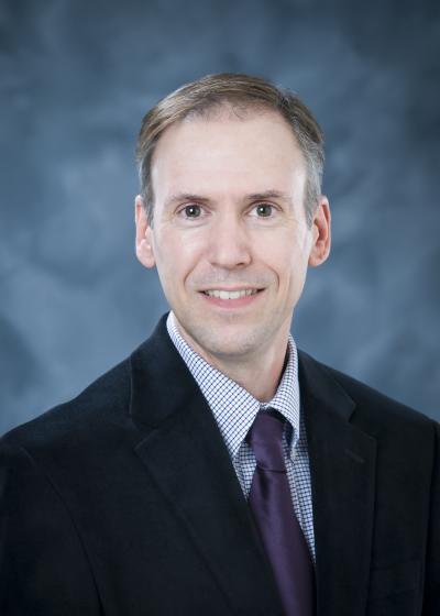 Professional photo of John Morris, wearing a black suit and purple tie against a blue background.