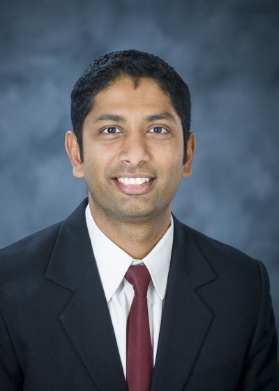 Professional photo of Varun Paul, wearing a black suit and red tie against a blue background.
