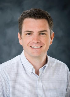 Professional photo of Adam Skarke wearing a white dress shirt against a gray background. 